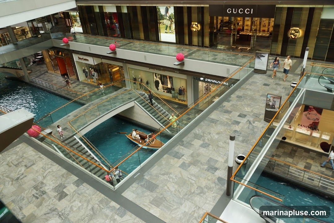 An aerial view of a modern indoor shopping mall featuring a canal with a boat and several luxury brand stores. The stores visible include Gucci, Juicy Couture, and Swarovski. People are seen walking on the elevated walkways and riding in the boat.