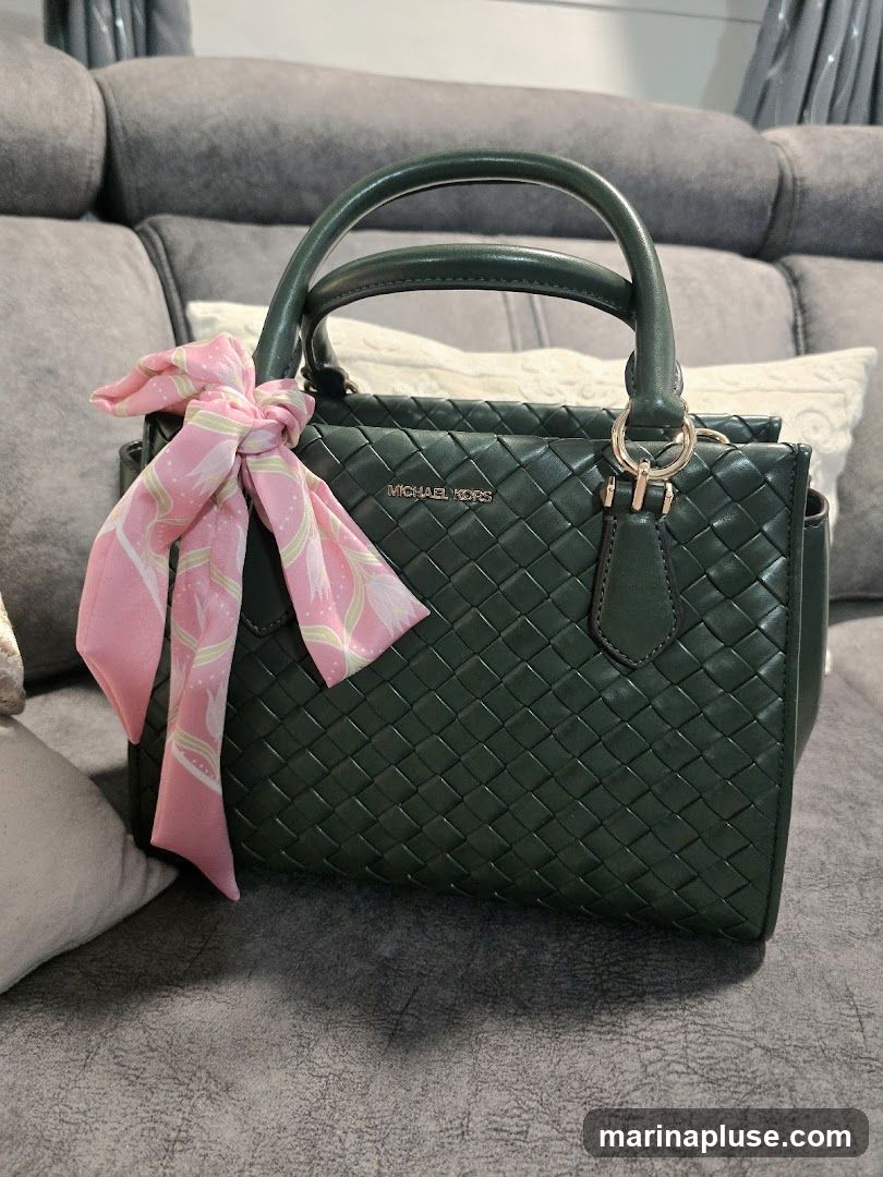 A dark green, woven leather handbag with a pink scarf tied around one of its handles. The Michael Kors logo is visible on the front of the bag. The bag is resting on a grey couch with pillows.