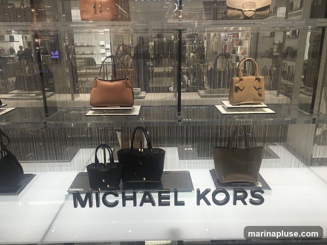 A display of various Michael Kors handbags in different sizes and colors, arranged on shelves within a store. The brand name 'MICHAEL KORS' is prominently displayed in large, black, three-dimensional letters at the bottom of the display. The background shows reflections of the store interior and other merchandise.
