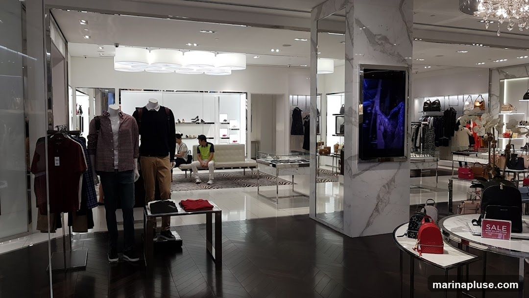 The image shows the interior of a modern fashion store. Mannequins are dressed in casual wear, and racks of clothing and handbags are displayed throughout the space. A digital screen on a marble-clad pillar shows a fashion-related advertisement. There are also display tables with accessories and a seating area for customers. A "SALE" sign is visible on a table in the foreground.
