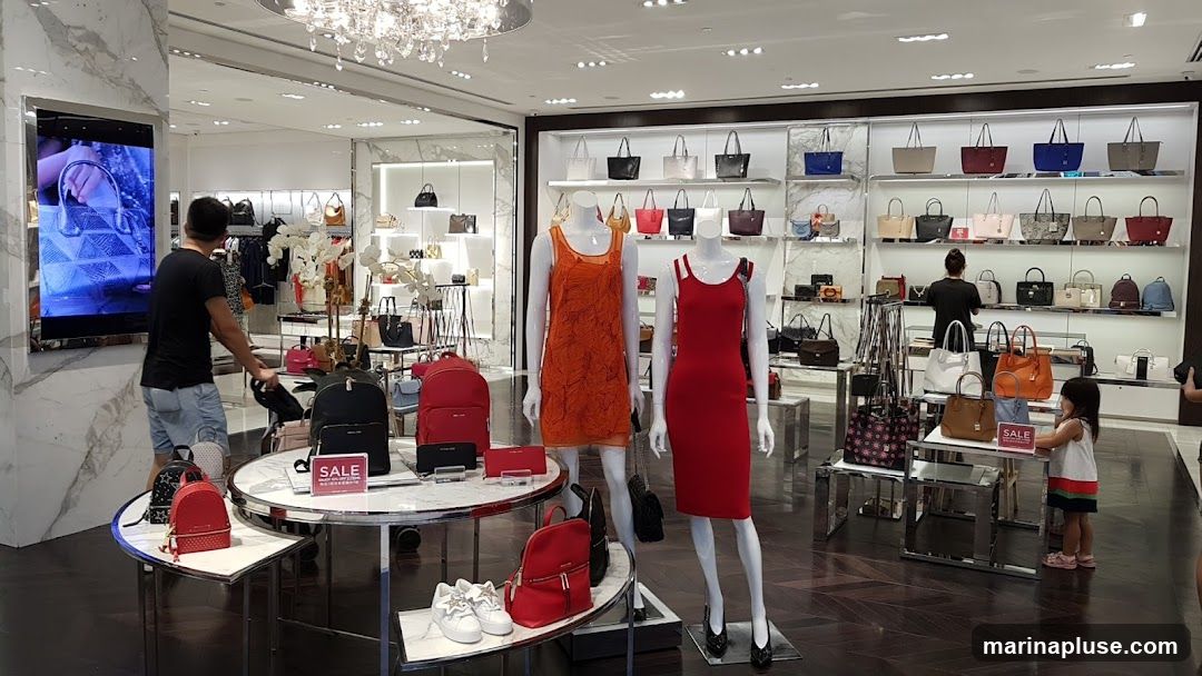 A well-lit interior of a store, likely a handbag boutique, showcasing a variety of handbags and accessories. Mannequins are dressed in colorful dresses, and displays feature handbags of different sizes, colors, and styles. A man is seen looking at a screen displaying a handbag, and a young child is interacting with a display stand. Signs indicating "SALE" are visible on some displays.