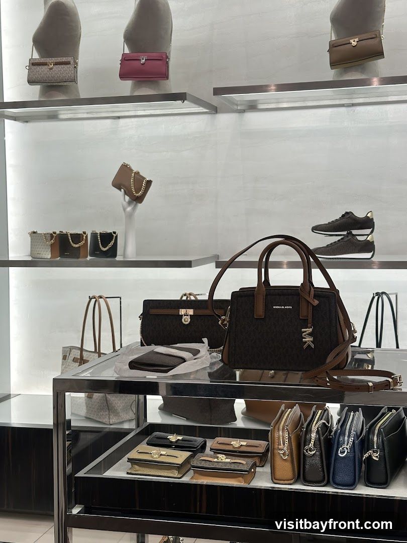 The interior of a high-end store displays a wide selection of handbags and accessories on illuminated shelves and racks. Mannequins are dressed in stylish outfits, showcasing the store's fashion offerings. The store features a modern and luxurious design with dark wood flooring and sleek white shelving.