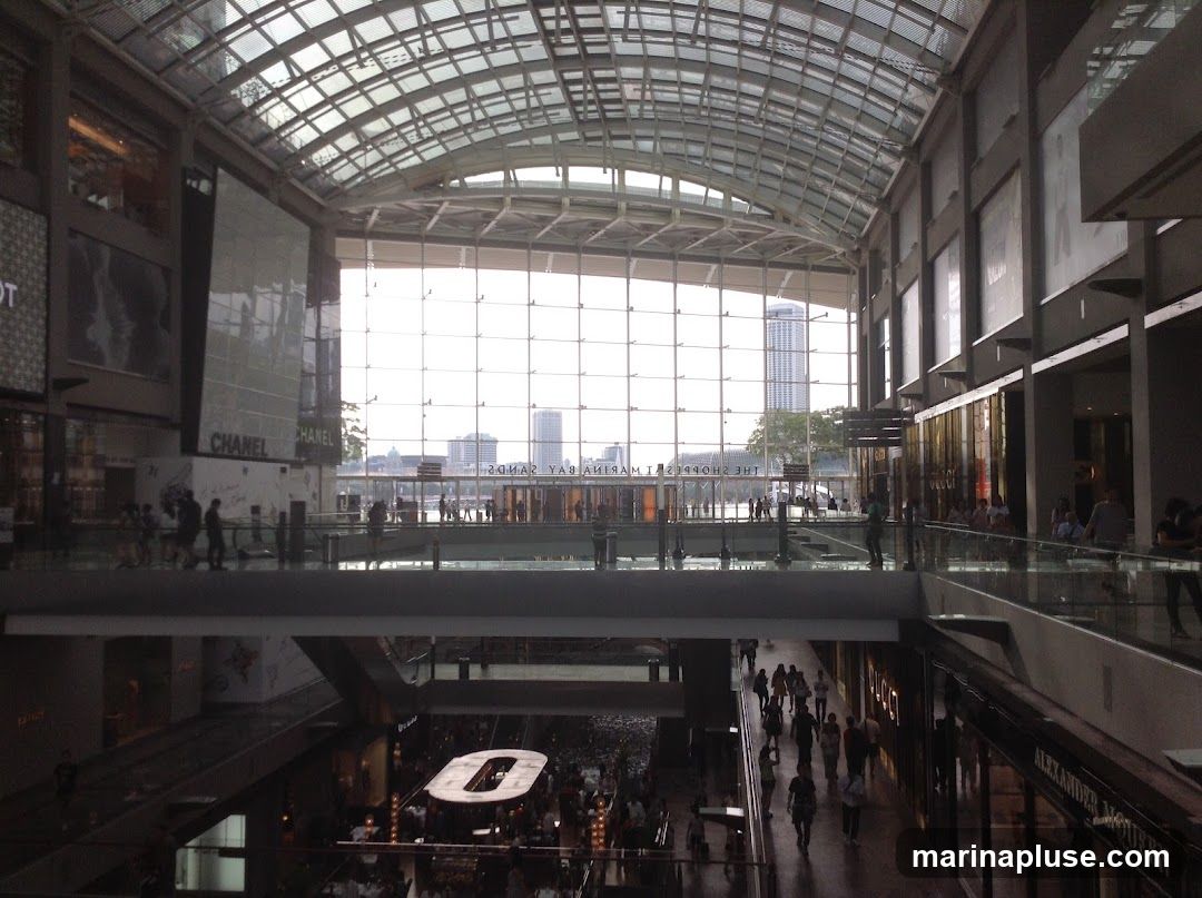 The image shows the interior of a modern shopping mall with a glass ceiling and large windows overlooking a cityscape. Multiple levels of shops and walkways are visible, with many shoppers browsing. Brands like Chanel and Alexander McQueen are advertised on storefronts.