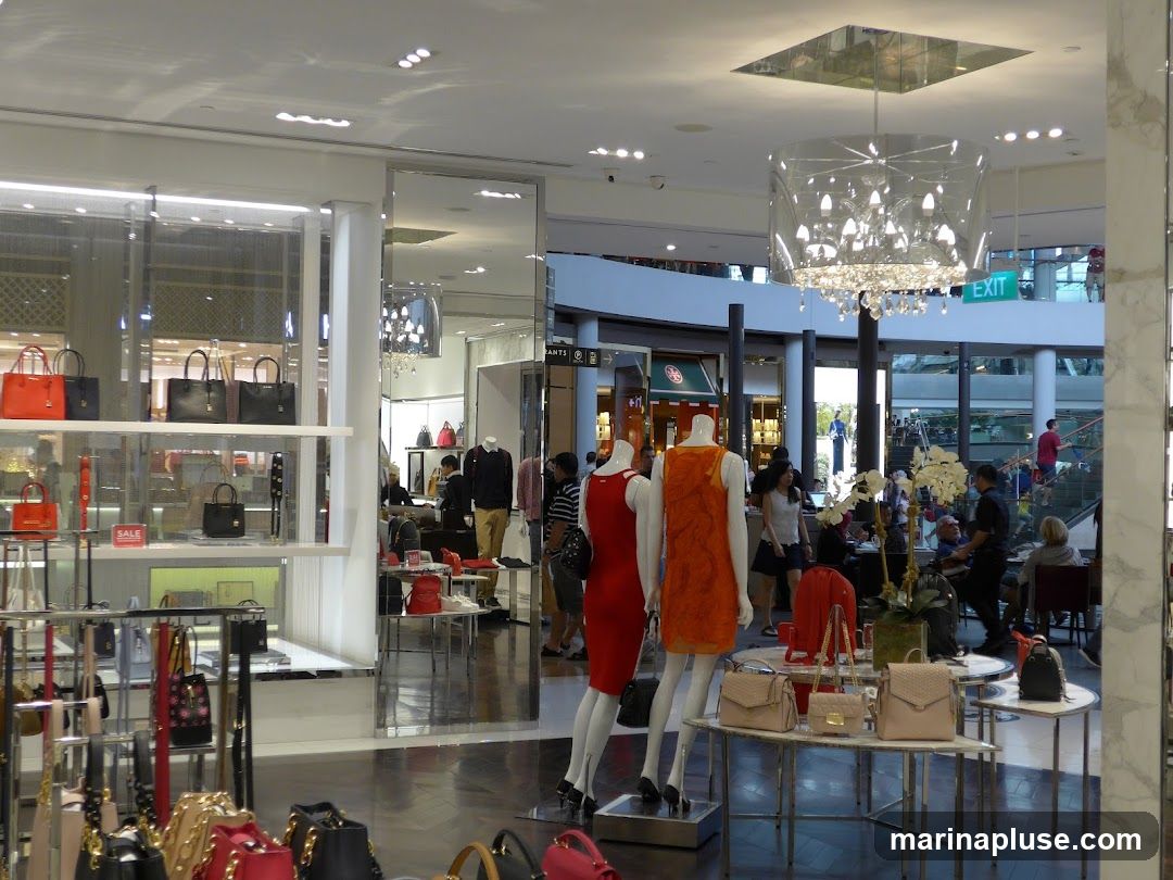 The image shows the interior of a brightly lit fashion store. Mannequins are displayed wearing dresses, and shelves are filled with handbags and accessories. In the background, other shoppers can be seen browsing and walking through the store. A large, ornate chandelier hangs from the ceiling, and an "EXIT" sign is visible.