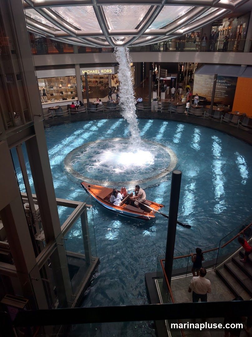 An indoor scene featuring a canal with a boat carrying passengers, a large waterfall cascading into the water, and surrounding shops and walkways. The architecture includes a modern, multi-level structure with a glass ceiling. A prominent sign for "MICHAEL KORS" is visible on one of the shops.