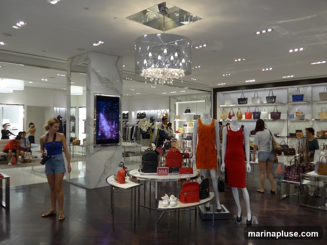 A well-lit, modern fashion store interior with displays of handbags, clothing on mannequins, and accessories. Several customers are browsing the merchandise. A large digital screen is mounted on a marble-clad pillar.