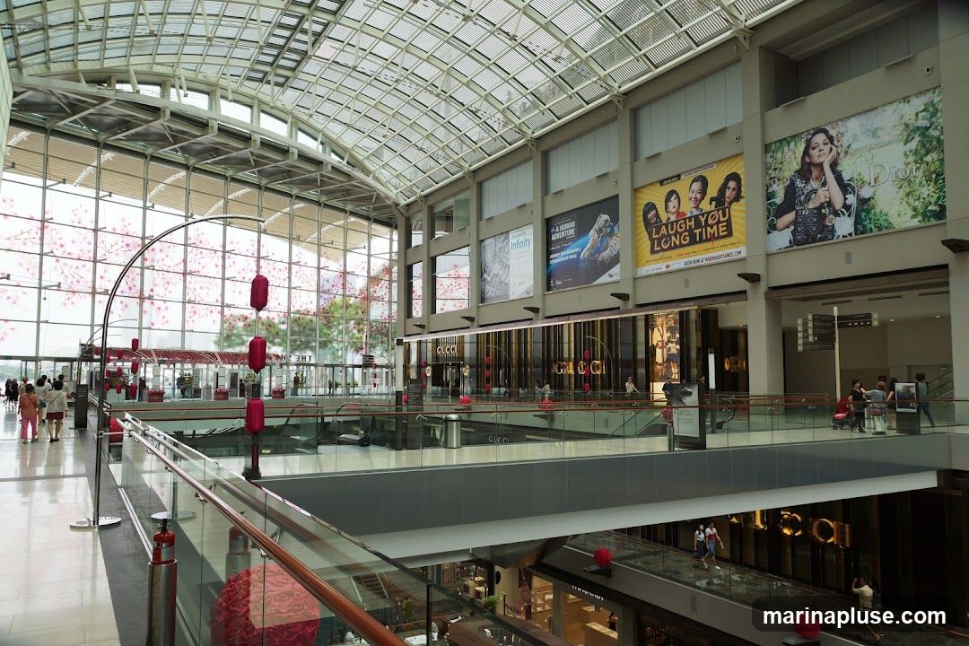 The image shows the interior of a multi-level shopping mall with a glass ceiling and large windows. Several shops are visible on different levels, including a prominent Gucci store. Large advertisements and posters are displayed on the walls. People are seen walking around and browsing. Decorative elements like lanterns and floral arrangements add to the ambiance.