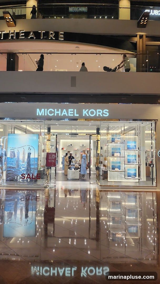 The exterior of a Michael Kors store with large glass windows and doors. The store's name is prominently displayed in illuminated letters above the entrance. Inside, mannequins display clothing and shelves are stocked with handbags and accessories. A large poster on the left window advertises a sale. The floor reflects the store's interior and signage.