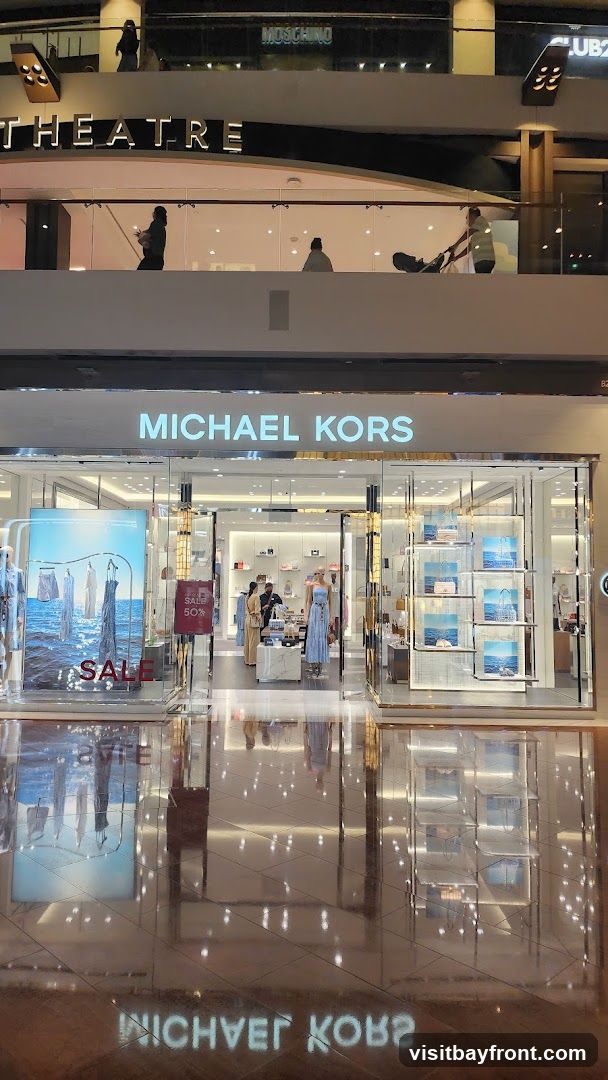 The exterior of a Michael Kors store with large glass windows and doors. The store's name is prominently displayed in illuminated letters above the entrance. Inside, mannequins display clothing and shelves are stocked with handbags and accessories. A large poster on the left window advertises a sale. The floor reflects the store's interior and signage.