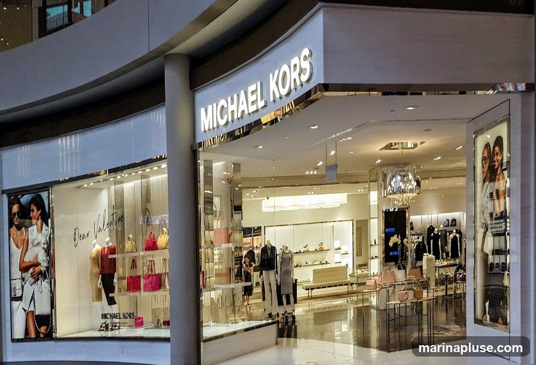 The exterior of a Michael Kors store with a large illuminated sign above the entrance. The storefront features large display windows showcasing handbags, clothing on mannequins, and promotional imagery. The interior of the store is visible, displaying more merchandise and a chandelier.