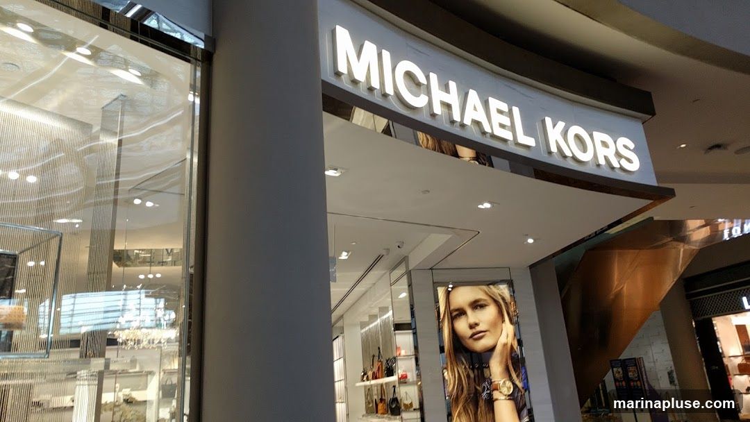The exterior of a Michael Kors store with illuminated signage above the entrance. A large advertisement featuring a woman wearing a watch is displayed to the right of the entrance. The store interior, visible through the glass facade, shows displays of handbags and accessories.