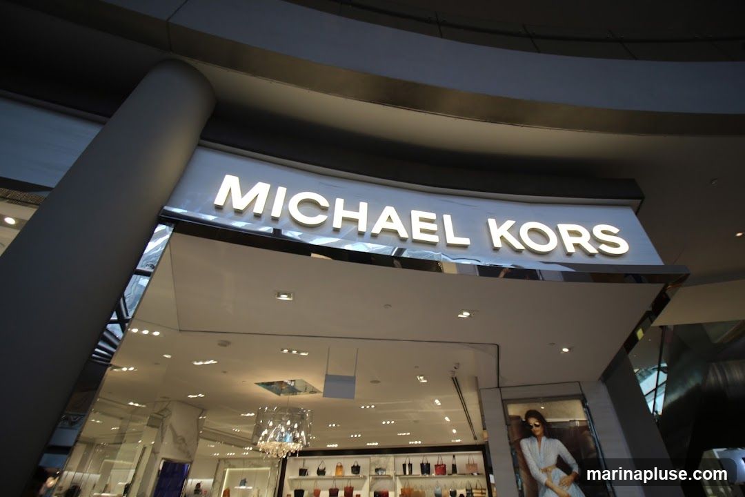 The exterior of a Michael Kors store with illuminated signage above the entrance. The store interior is visible through the large glass windows, displaying handbags and other merchandise. A promotional image of a woman is also visible to the right of the entrance.