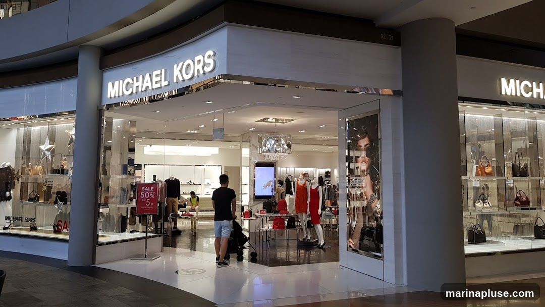 The exterior of a Michael Kors store with large glass windows displaying handbags and clothing. The store's name is prominently displayed in illuminated letters above the entrance. A sale sign is visible in the window.
