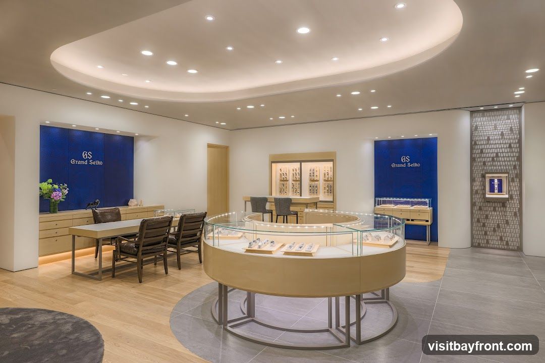 The interior of a Grand Seiko watch store features a circular display case in the center, showcasing watches. To the left, a wooden counter with chairs is set up, and behind it, a blue wall displays the Grand Seiko logo. To the right, another display case and a wall-mounted advertisement are visible. The store has a modern and luxurious design with wooden floors and a curved ceiling with recessed lighting.