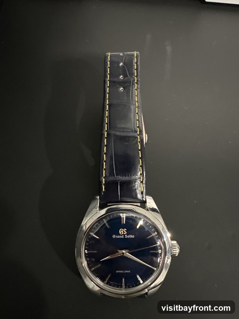 A close-up, top-down view of a Grand Seiko Spring Drive watch with a dark blue dial and a dark blue alligator leather strap with yellow stitching. The watch has a silver-toned case and hands. The dial features the Grand Seiko logo and "SPRING DRIVE" text.