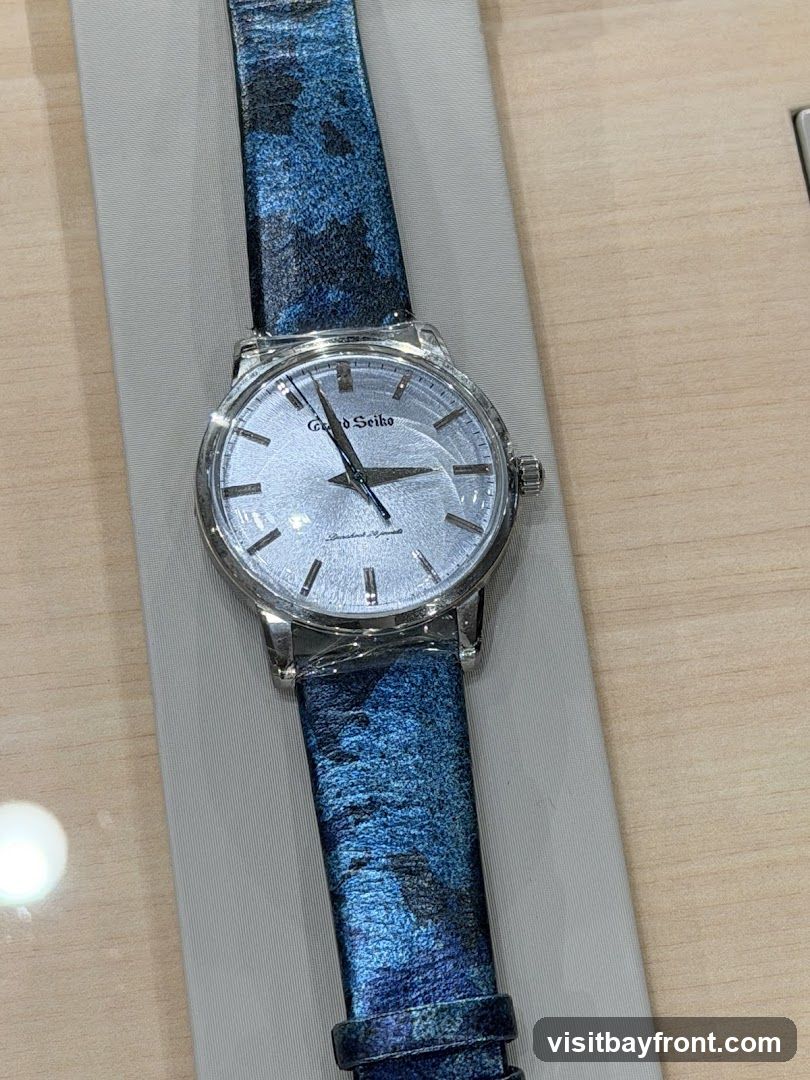 A close-up shot of a Grand Seiko watch with a silver dial and a blue patterned strap. The watch is displayed on a light gray fabric surface, with a wooden surface visible in the background.