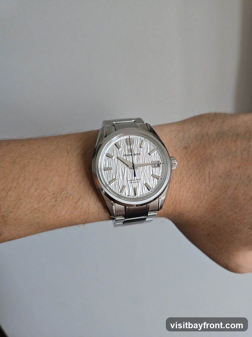 A close-up shot of a Grand Seiko watch with a textured silver dial and a metal bracelet, worn on a person's wrist against a plain background.