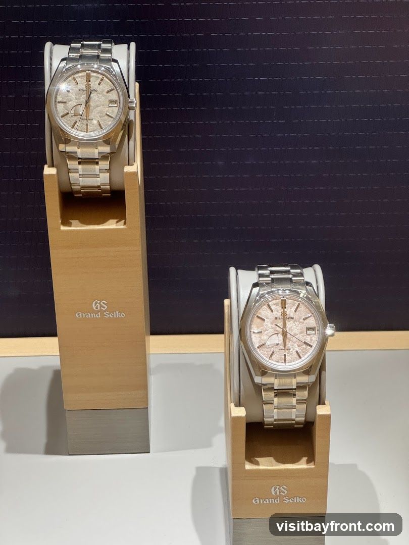 Two Grand Seiko watches are displayed on wooden stands against a dark, textured background. The watches have silver metal bands and light-colored, textured dials. The stands have the "GS Grand Seiko" logo printed on them.