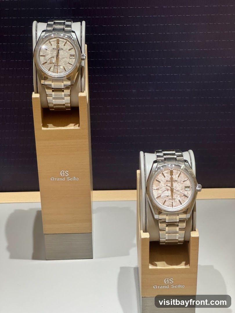 Two Grand Seiko watches are displayed on wooden stands. The watches have silver metal bands and light-colored textured dials. The stands have the Grand Seiko logo printed on them. The background is dark with a subtle striped pattern.