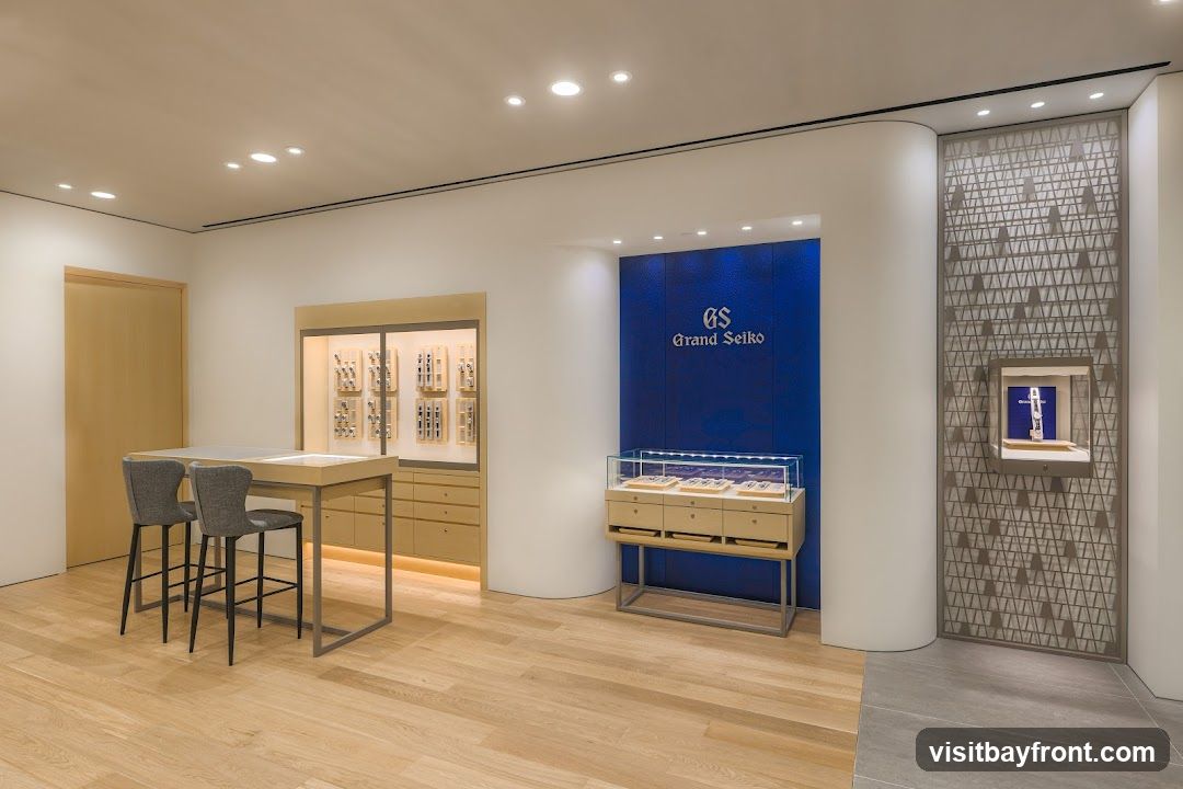 A modern and minimalist interior of a Grand Seiko watch store. The space features light wood flooring, white walls, and subtle lighting. Display cases showcase various watches. A prominent blue wall with the Grand Seiko logo is visible. There is a consultation table with two chairs.