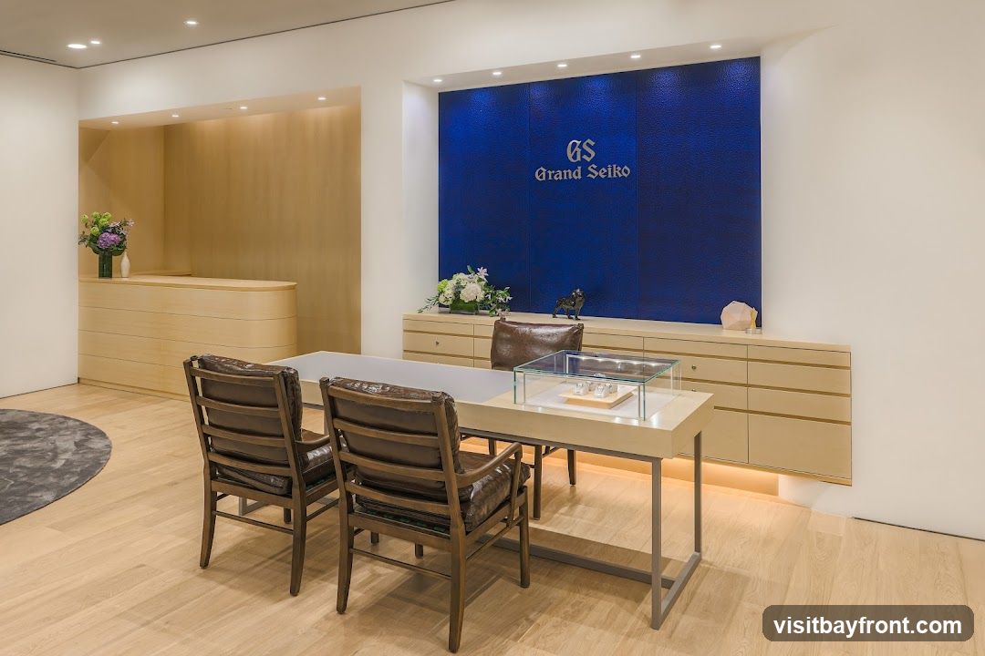 A well-lit showroom with a blue textured wall featuring the Grand Seiko logo. A light wood reception desk is visible on the left, and a display table with chairs is in the foreground. A glass display case on the table showcases watches.