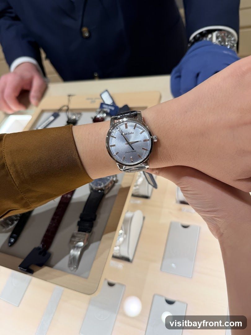 A person is trying on a silver watch with a light blue textured dial. The watch has a metal band and the brand name "Grand Seiko" is visible on the dial. In the background, a person in a dark suit and blue gloves is visible, along with other watches and display stands on a wooden counter.