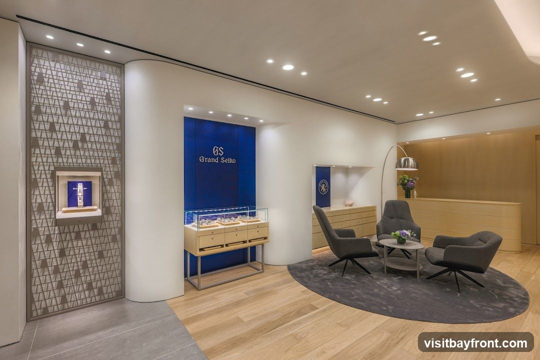 The interior of a Grand Seiko watch store features a display of watches in a glass case, a decorative wall panel with a watch display, and a seating area with armchairs and a coffee table. A reception desk is visible in the background.