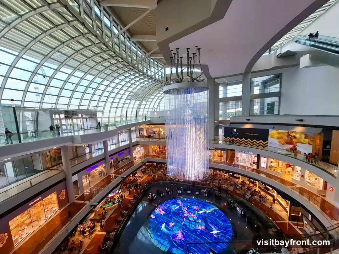 The image shows the interior of a multi-level shopping mall. A large, cylindrical chandelier hangs in the center of the atrium, and a circular LED screen at the bottom displays a vibrant blue aquatic scene. Shops are visible on multiple floors, with escalators and walkways connecting them. People are seen browsing in shops and dining at tables. The mall has a modern design with a glass roof allowing natural light to enter.