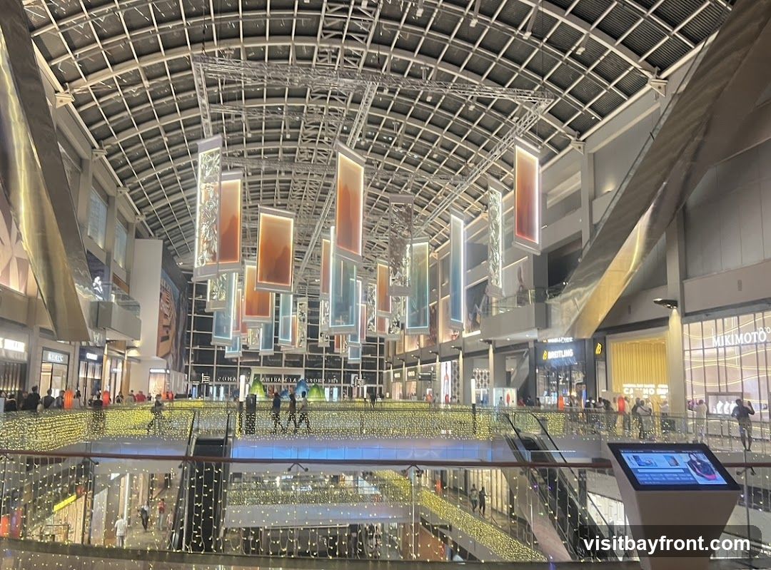 The image shows the interior of a large, modern shopping mall. The ceiling is a complex grid structure, and numerous rectangular art installations with colored lighting hang from the ceiling. The mall has multiple levels, with escalators visible. Balconies and walkways are adorned with strings of warm white fairy lights. People are seen walking on the different levels. Storefronts with illuminated signage are visible on the upper levels, including 'BREITLING' and 'MIKIMOTO'. A digital information kiosk is present in the foreground.