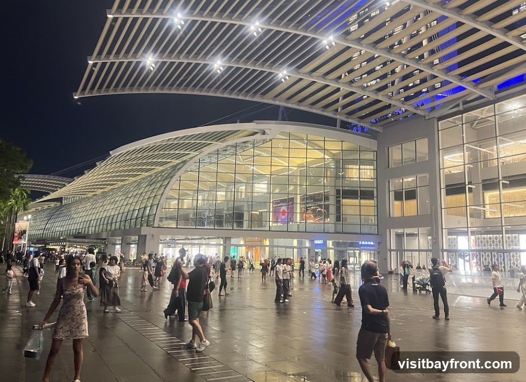 A wide shot of a contemporary shopping complex at night. The building features a large, curved glass facade illuminated from within, with a distinctive slatted roof structure overhead. Many people are walking in the open plaza area in front of the complex. The lighting creates a vibrant atmosphere.