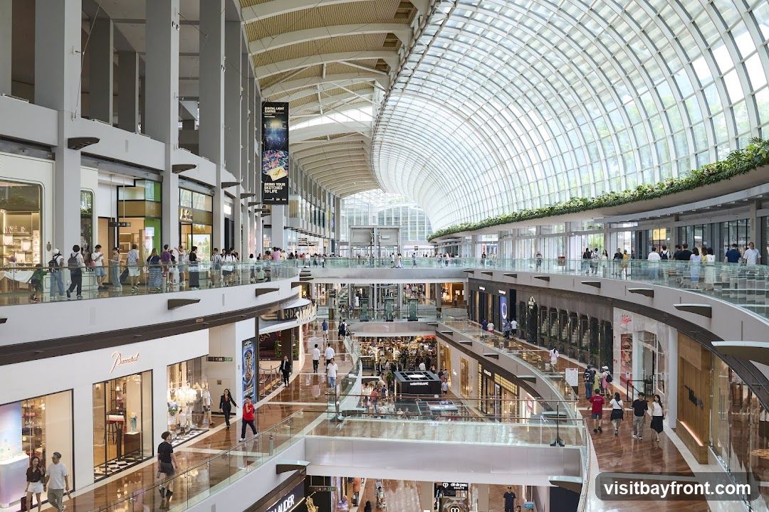 A wide-angle view of the interior of a large, multi-level shopping mall. The architecture features a curved glass ceiling, allowing natural light to flood the space. Multiple levels of shops and walkways are visible, with shoppers browsing and walking around. Some shopfronts are clearly visible, including one with the name 'Baccarat'. Banners hang from the ceiling, one of which reads 'BRING YOUR SKETCHES TO LIFE'. The overall impression is one of a bright, spacious, and upscale retail environment.