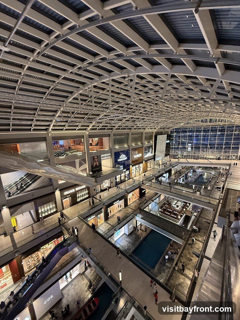 An aerial view of the interior of a modern shopping mall. The mall features multiple levels with escalators and walkways connecting different sections. Shops with visible signage are present on each level. In the center of the mall, there is a water feature and seating areas. The ceiling is a complex structure of metal beams and panels, with lighting illuminating the space. People are seen walking around and shopping.