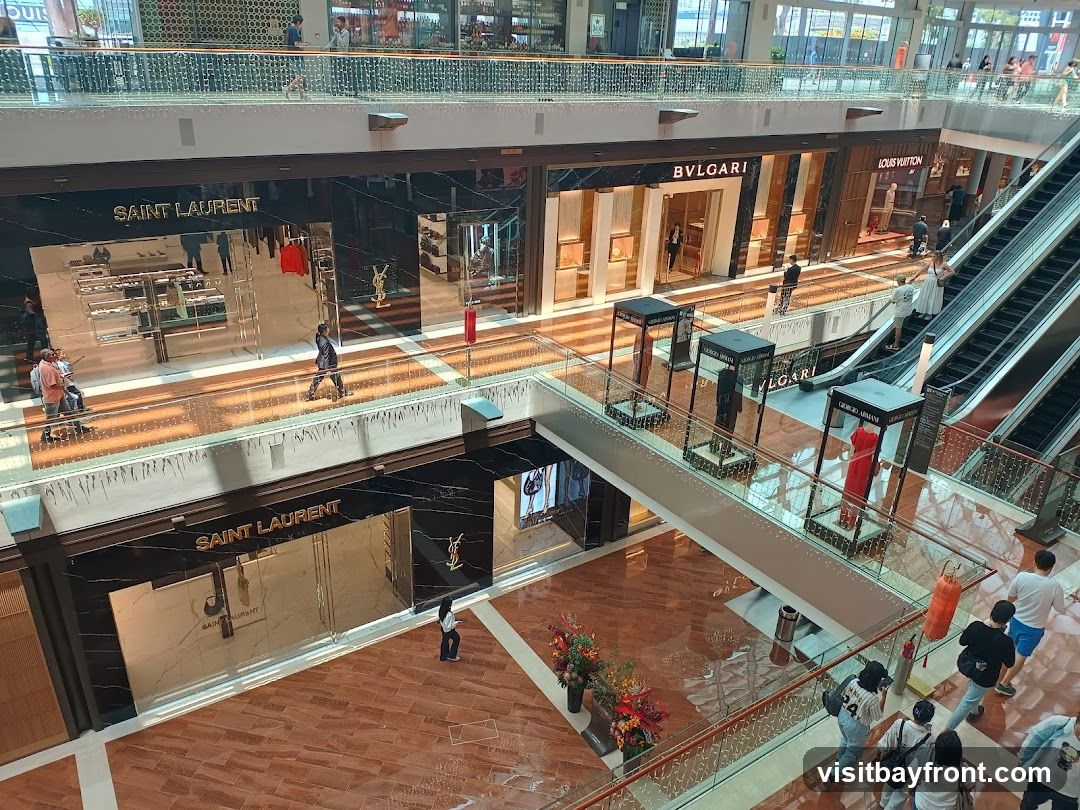 An interior view of a multi-level luxury shopping mall. Several high-end stores, including Saint Laurent, Bvlgari, and Louis Vuitton, are visible on the ground floor. Escalators ascend to upper levels where more shops and shoppers can be seen. Displays of merchandise and mannequins are present outside some of the stores.