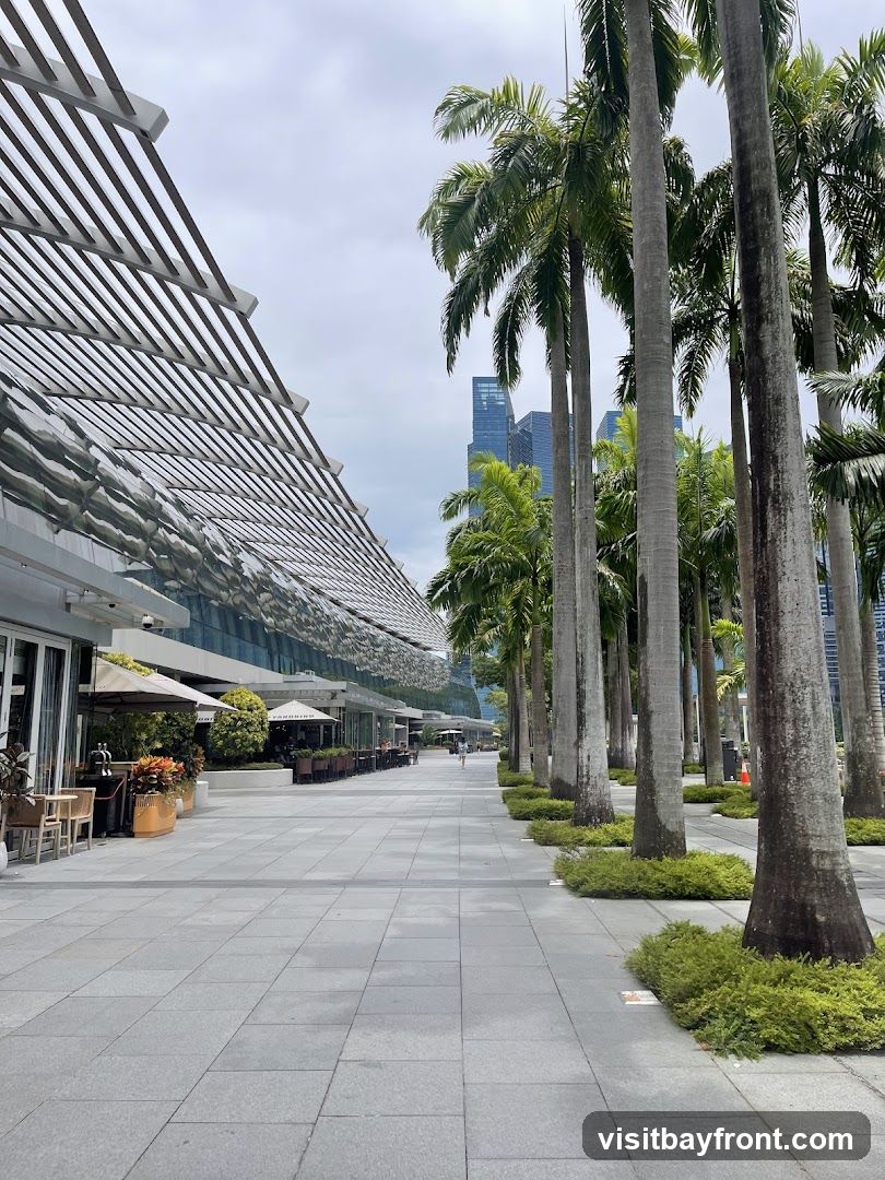 A wide, paved walkway lined with tall palm trees on the right. On the left, a modern building with a distinctive slatted roof extends into the distance. Several outdoor seating areas with umbrellas are visible in front of establishments along the building. In the background, modern skyscrapers rise against a cloudy sky.