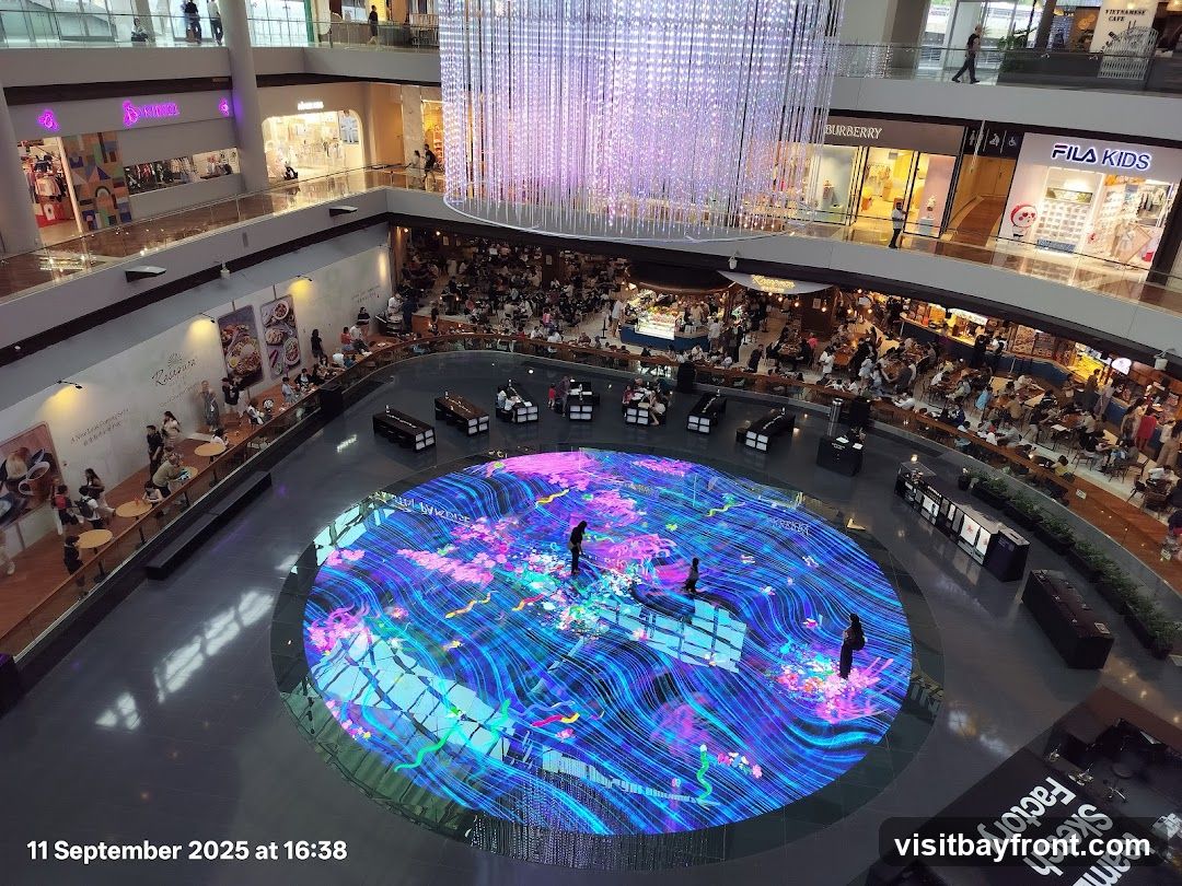 An overhead view of a bustling shopping mall atrium. A large, circular digital display on the floor shows a vibrant, abstract pattern of blues, purples, and pinks, with several people standing on it. Surrounding the atrium are multiple levels of shops, including visible storefronts for Burberry and Fila Kids. A large, ornate chandelier hangs from the ceiling. People are seen browsing shops and dining at various restaurants and cafes on different levels. The timestamp "11 September 2025 at 16:38" is visible in the bottom left corner.