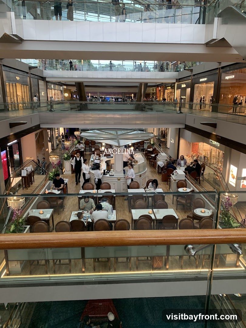 An overhead view of a busy shopping mall interior. Multiple levels of shops and walkways are visible, with shoppers browsing. In the foreground, a cafe with tables and chairs is set up, with staff serving customers. The cafe has a prominent sign that reads "ANGELINA".