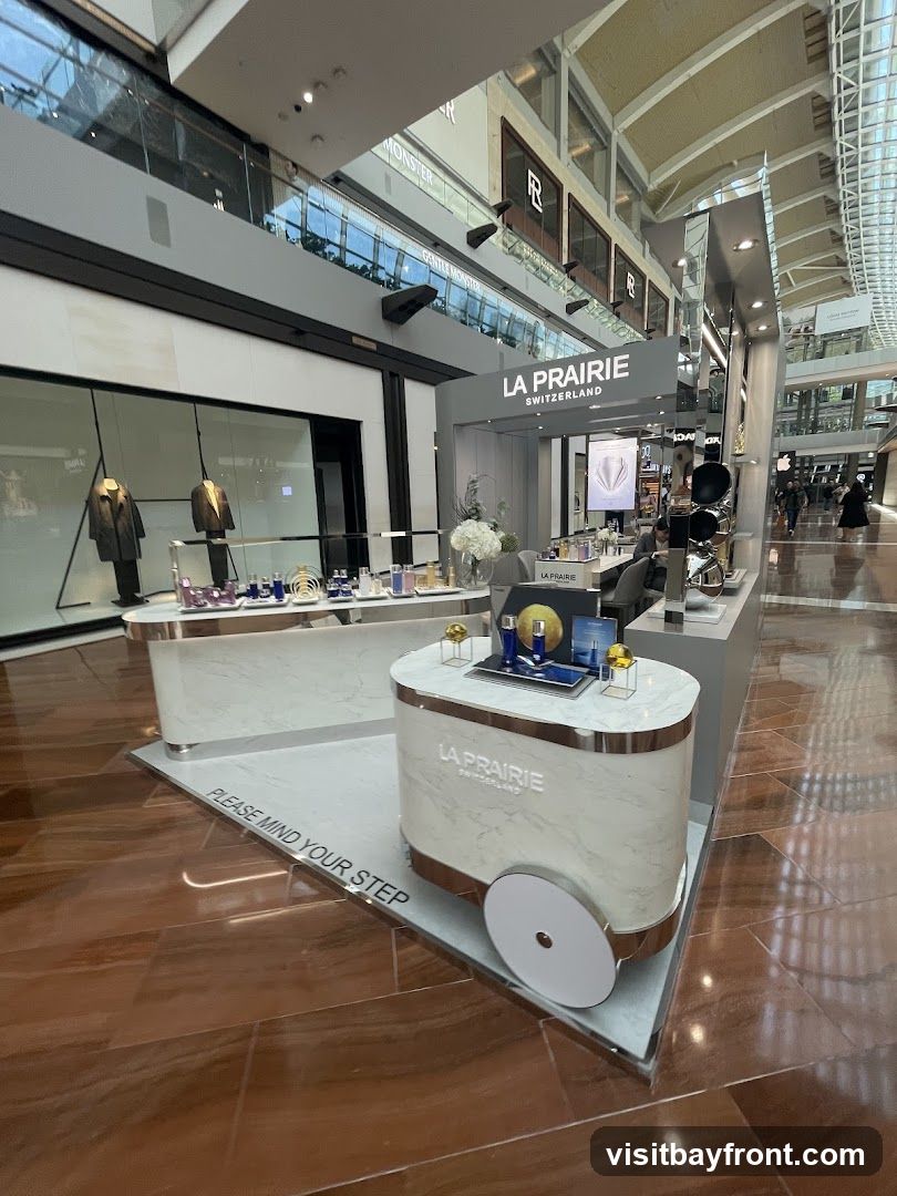 A La Prairie kiosk is set up in a shopping mall. The kiosk features marble-patterned displays with rose gold accents. Products are displayed on the counter, including bottles and jars of skincare. A sign above the kiosk reads "LA PRAIRIE SWITZERLAND." In the background, other shops and shoppers are visible.
