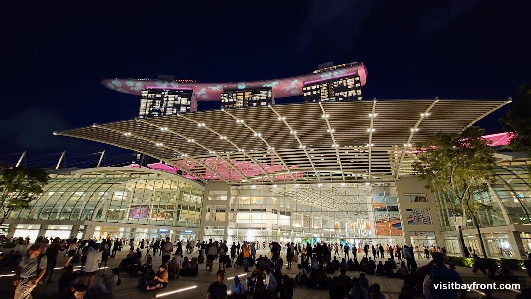 A wide shot shows a large, modern building complex at night, illuminated by lights. The building features a distinctive curved roof structure and extensive glass facades. In the background, a taller building is visible with illuminated patterns on its exterior. A large crowd of people are gathered in the open area in front of the complex, many sitting on the ground. The scene suggests a public gathering or event.