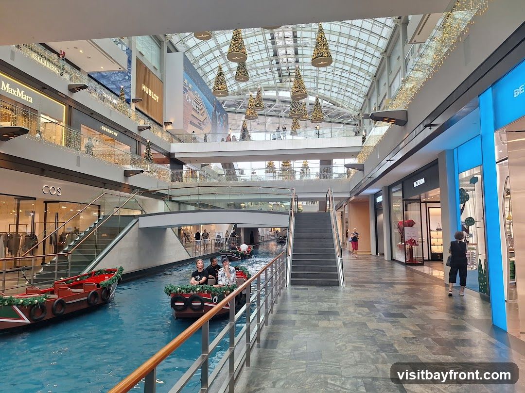 An indoor shopping mall features a canal with gondolas carrying passengers. Shops like MaxMara, Givenchy, Hublot, and COS are visible on multiple levels. The mall has a modern design with a glass ceiling and festive decorations, including Christmas trees and lights.