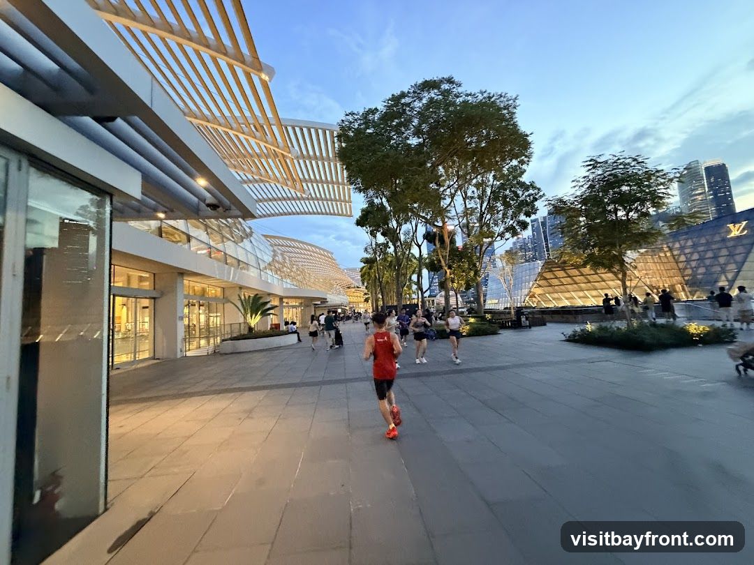 A wide paved walkway with people jogging and walking. On the left, there is a modern building with large glass windows and a white slatted roof structure. On the right, a distinctive building with a large 'LV' logo is visible, illuminated against the twilight sky. Trees line the walkway, and city skyscrapers are in the background.