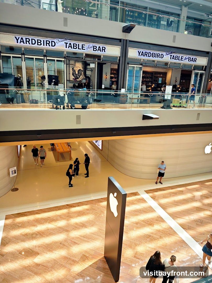 The image shows the interior of a modern shopping mall. On the upper level, two storefronts for "YARDBIRD Southern TABLE And BAR" are visible, with people inside. Below, an Apple Store is depicted with shoppers browsing and an Apple logo sign in the foreground. Sunlight streams through the ceiling, creating patterns on the floor.