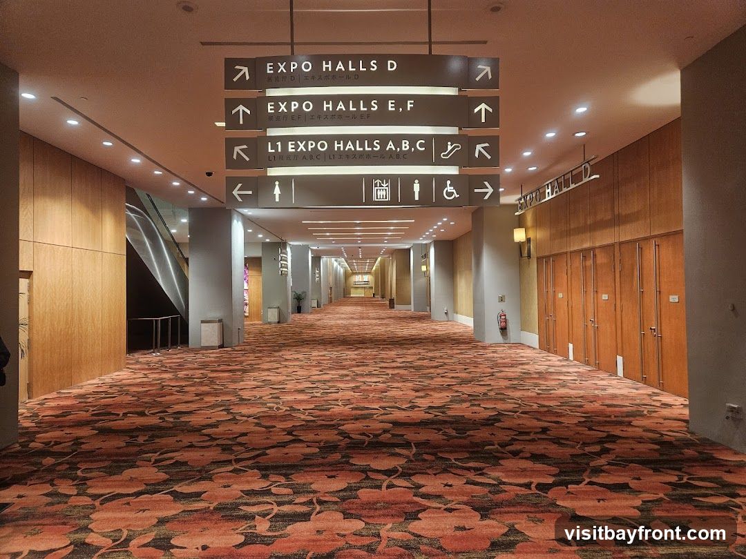 A long, wide hallway in a convention center with a patterned carpet. Overhead signs direct visitors to various expo halls and levels. Escalators and wooden doors are visible along the walls.
