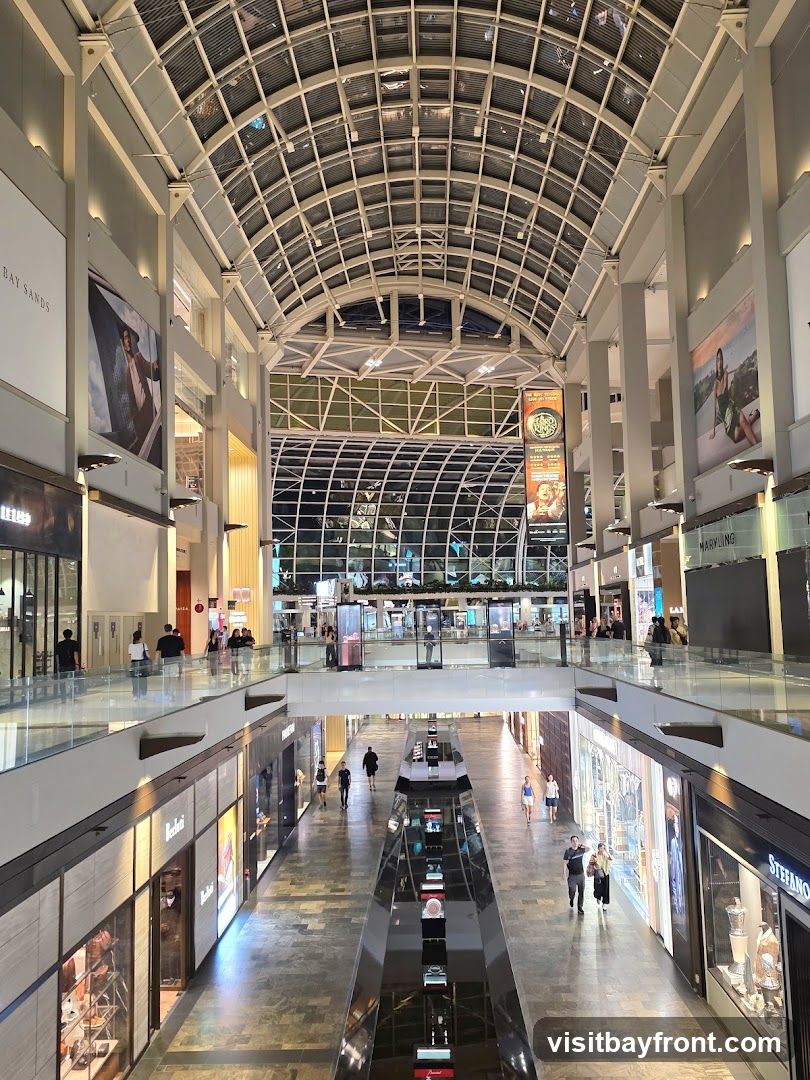 A wide-angle view of the interior of a large, modern shopping mall. The image looks down a long corridor with shops on either side and a reflective water feature in the center. The ceiling is a complex glass and metal structure, allowing natural light to filter in. People are seen walking along the mall, browsing in shops, and admiring the displays. Large advertisements and store signage are visible on the walls and storefronts.