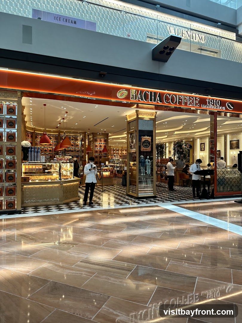 The exterior of a Bacha Coffee shop with its prominent orange signage and ornate gold detailing. Inside, displays of coffee products and pastries are visible, along with staff attending to customers. The shop is located within a modern shopping mall, with other stores like Valentino visible in the background.