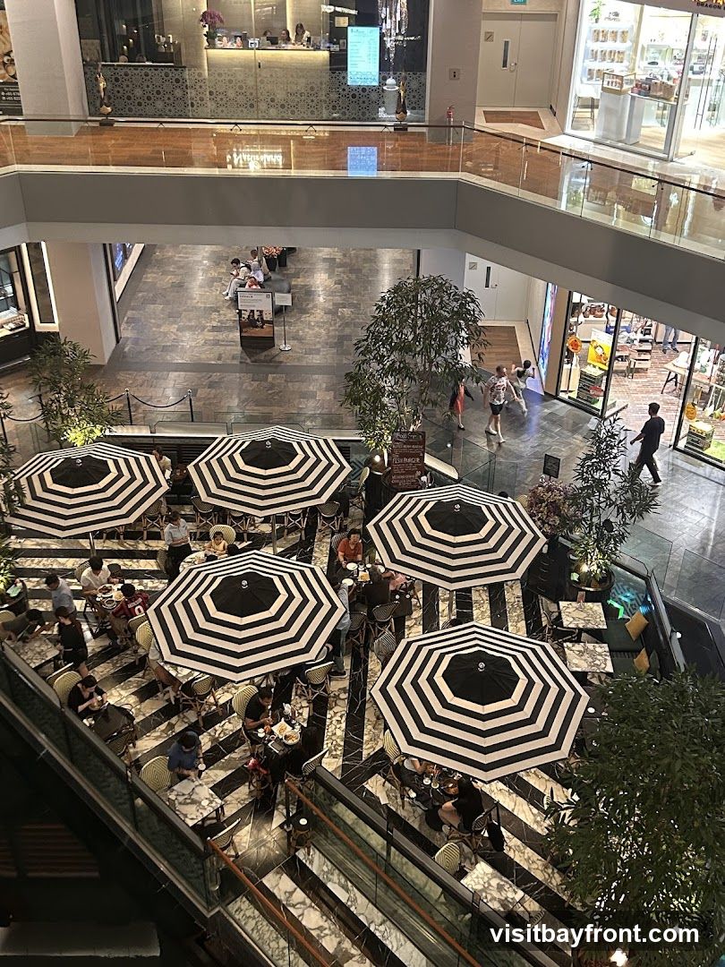 An overhead view of an outdoor cafe seating area within a multi-level shopping mall. The cafe features numerous black and white striped umbrellas providing shade over tables where patrons are seated. The floor has a distinctive black and white marble pattern. In the background, other shops and mall walkways are visible, with people browsing and walking.