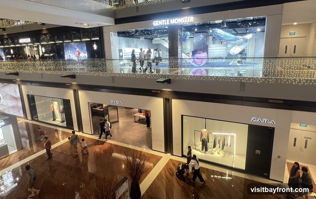 An interior view of a multi-level shopping mall with various stores visible. On the upper level, a store with the sign "GENTLE MONSTER" is prominent, featuring a modern display with artistic elements. Below, several "ZARA" stores are visible, showcasing clothing on mannequins and racks. The mall has a polished wooden floor and is decorated with string lights. People are seen walking around and browsing the shops.