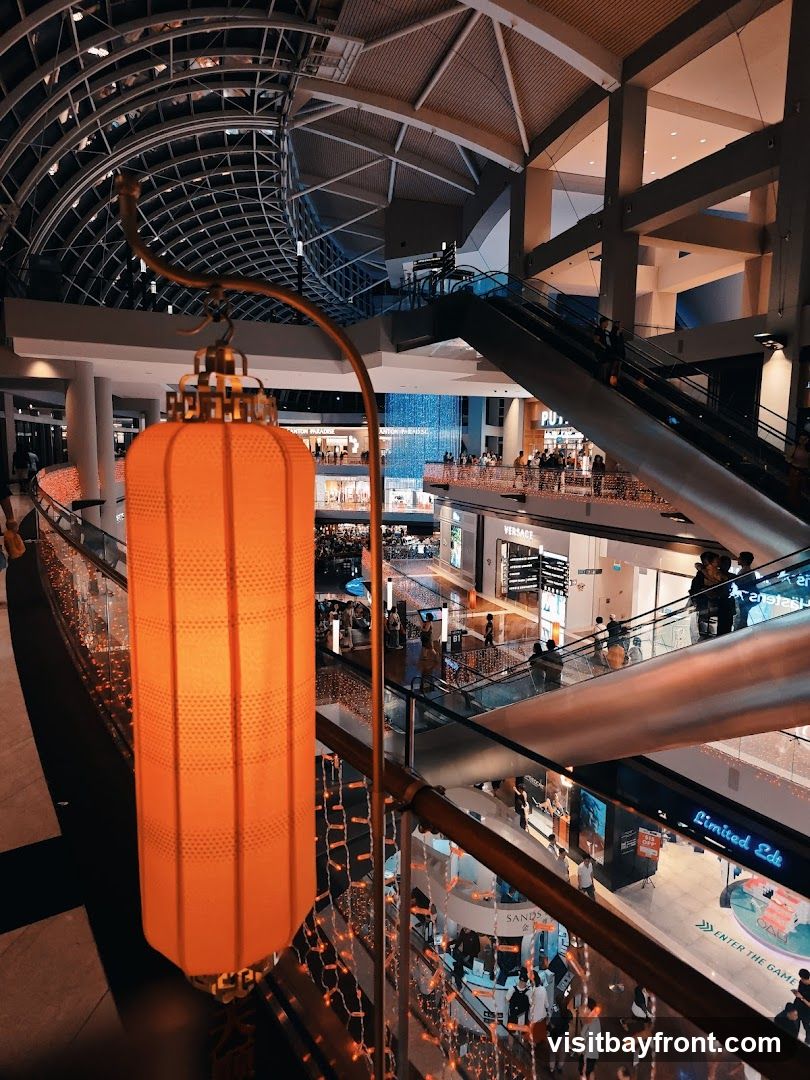 The image shows the interior of a multi-level shopping mall. In the foreground, a large orange lantern hangs from a decorative stand, adorned with small fairy lights. Escalators are visible, with people moving between floors. Various shop fronts are seen on different levels, some with illuminated signage. A large, abstract water feature or light installation is visible in the background. The architecture features a modern design with a domed ceiling and exposed structural elements.