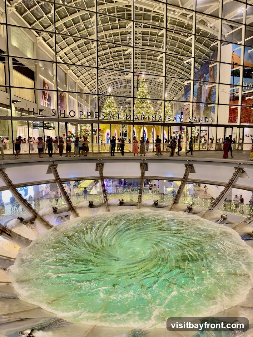 A view of the interior of a large shopping mall, featuring a large vortex fountain in the foreground. In the background, the text "THE SHOPPES AT MARINA BAY SANDS" is visible on the facade of the building, with Christmas trees and festive lights decorating the space. People are seen walking around the mall.
