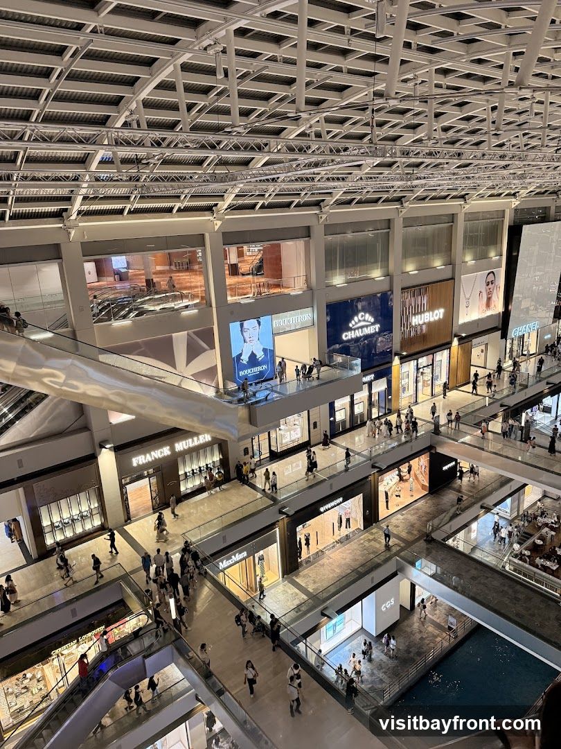 An aerial view of the interior of a modern, multi-level shopping mall. The image showcases various luxury brand stores with prominent signage, including Boucheron, Chaumet, Hublot, Chanel, Franck Muller, and MaxMara. Escalators and walkways connect different levels, and shoppers are seen browsing the stores and moving between levels. The mall features a contemporary architectural design with a high ceiling and intricate metal framework. A water feature is visible on one of the lower levels.
