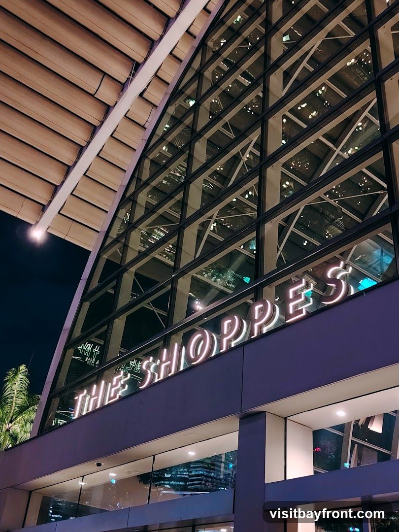 A neon sign spelling out "THE SHOPPES" is mounted on the exterior of a modern building with a large glass facade. The building has a distinctive angled roof structure.